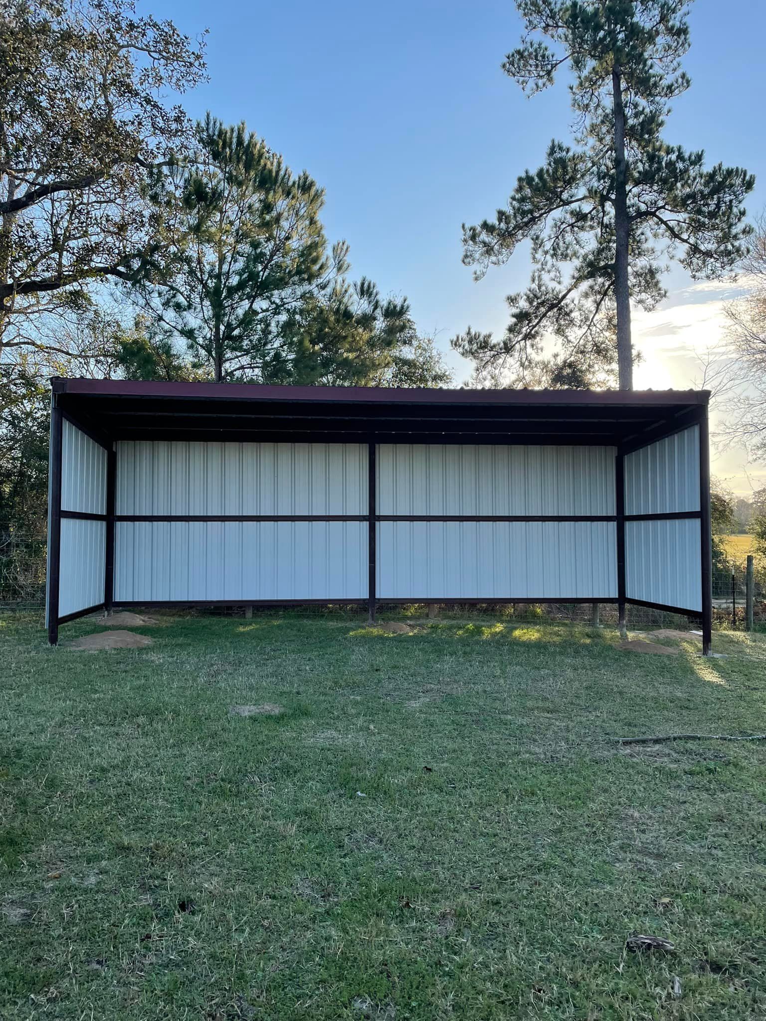 Open-air shelter with a maroon roof and white corrugated metal siding on a grassy field, trees in the background.