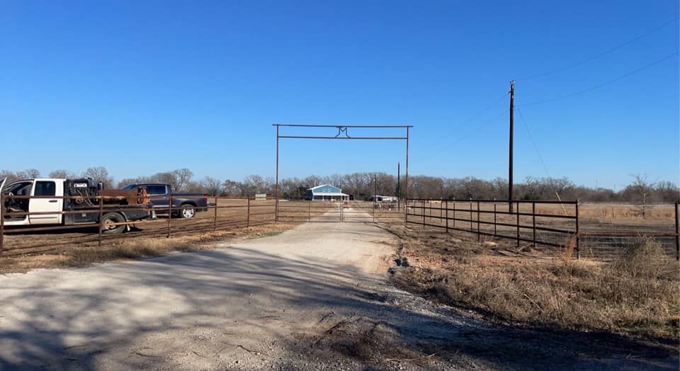 A rural gate leads to a ranch. A truck and trailer are parked to the left. Blue sky.