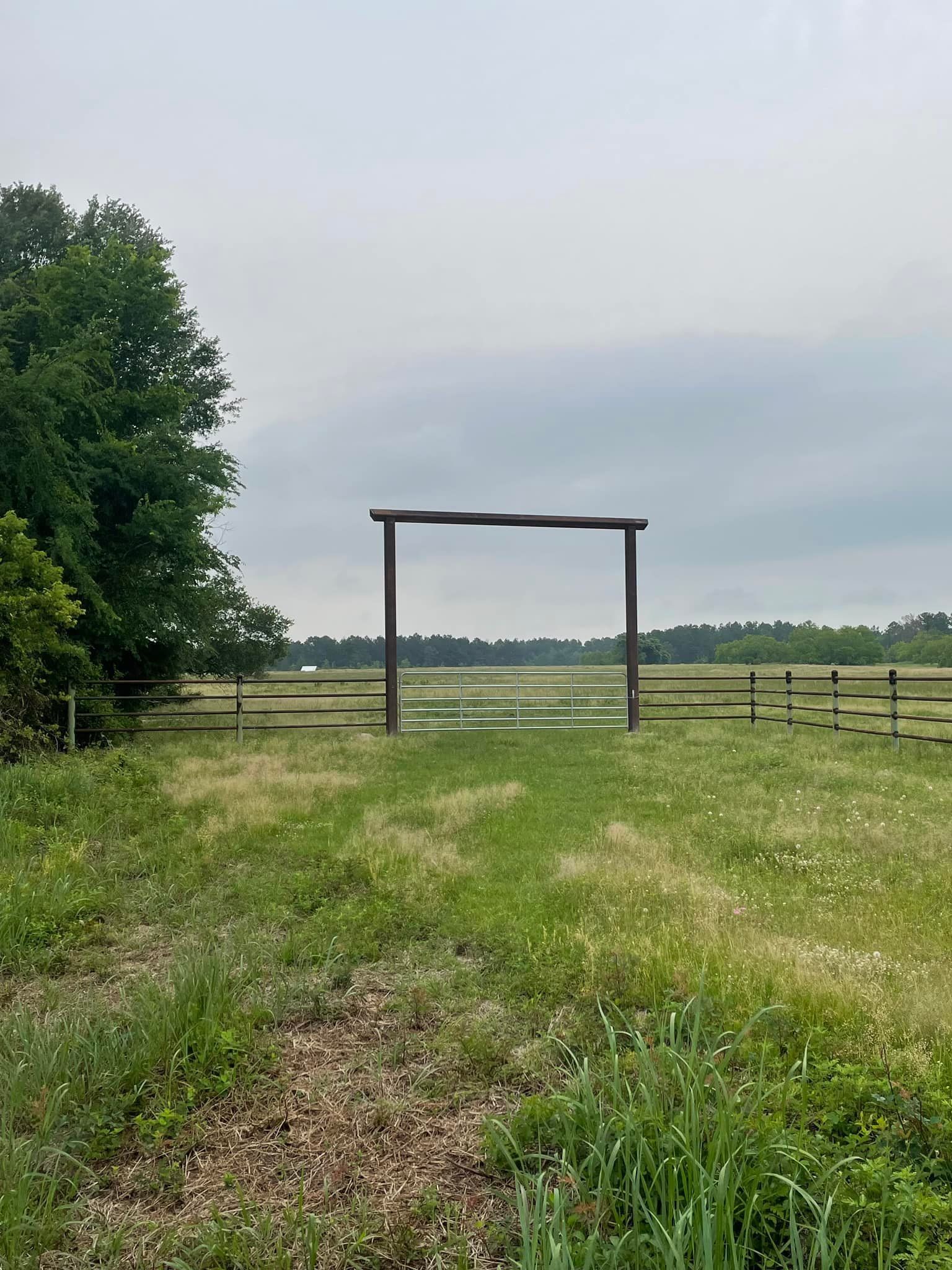 Wooden gate in a green field, with a fence and trees. Cloudy sky in the background.