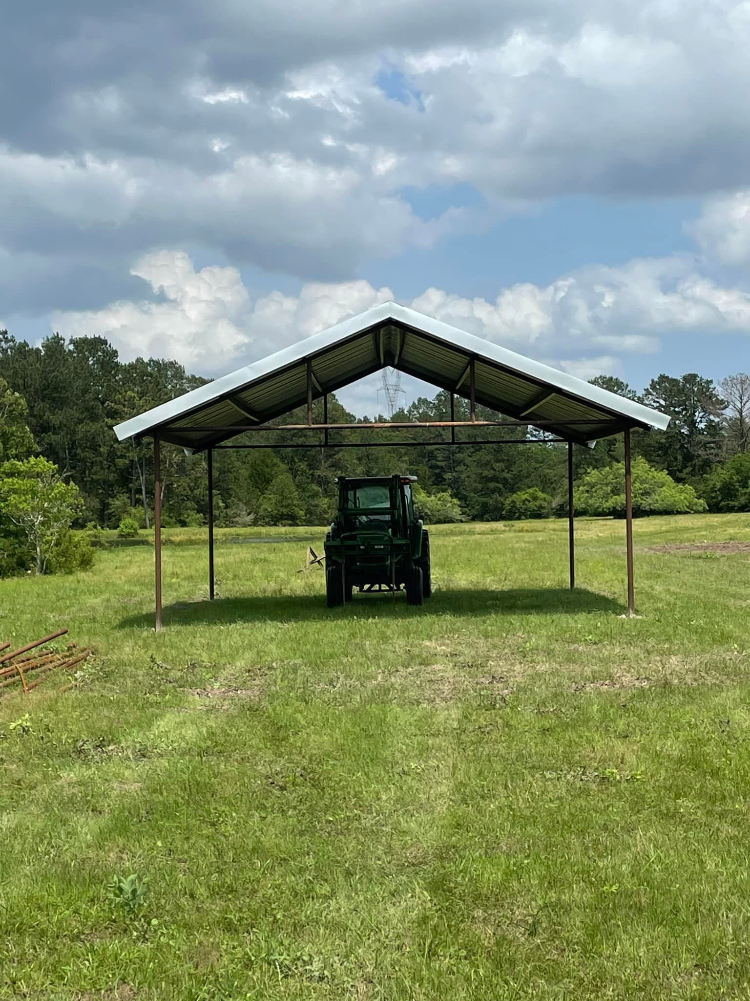 Green tractor under a metal roof carport in a grassy field on a cloudy day.