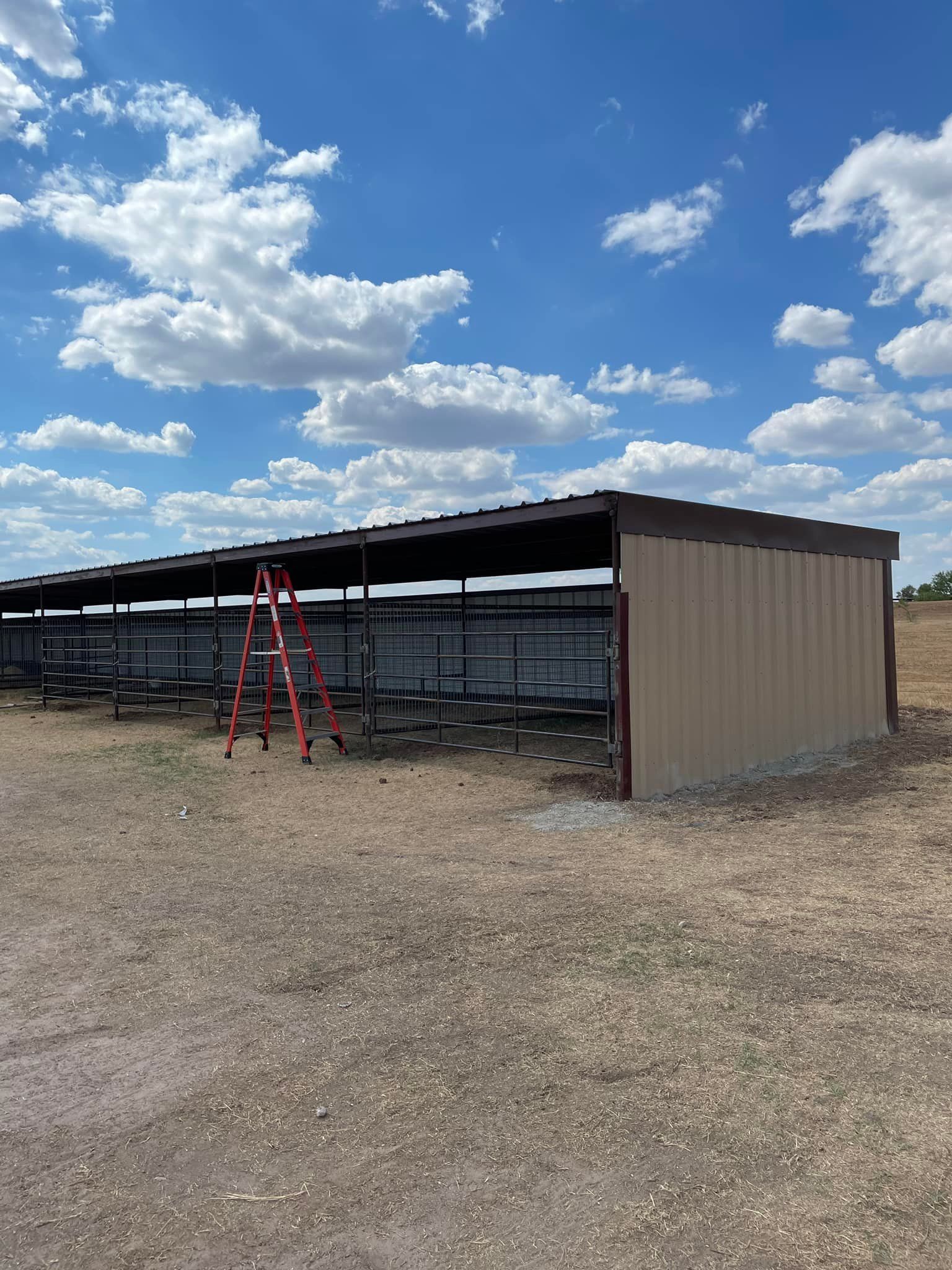 Tan and brown metal shelter under a bright blue sky with fluffy clouds; a red ladder is leaning against it.