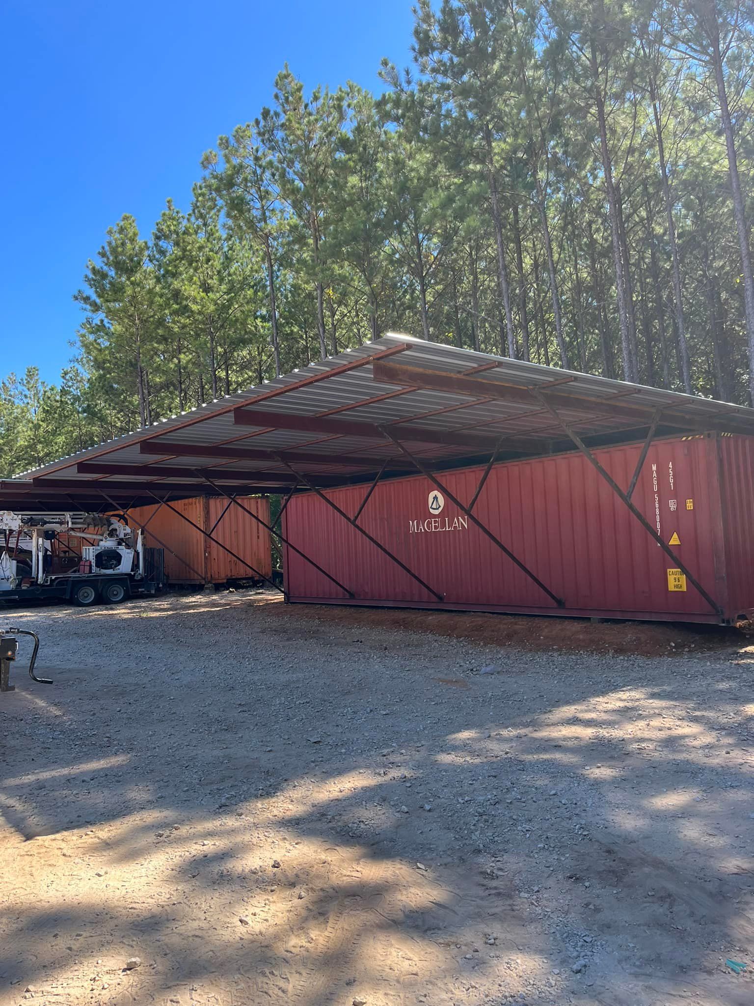Red shipping containers with metal roofs, in a gravel lot, with trees in background.