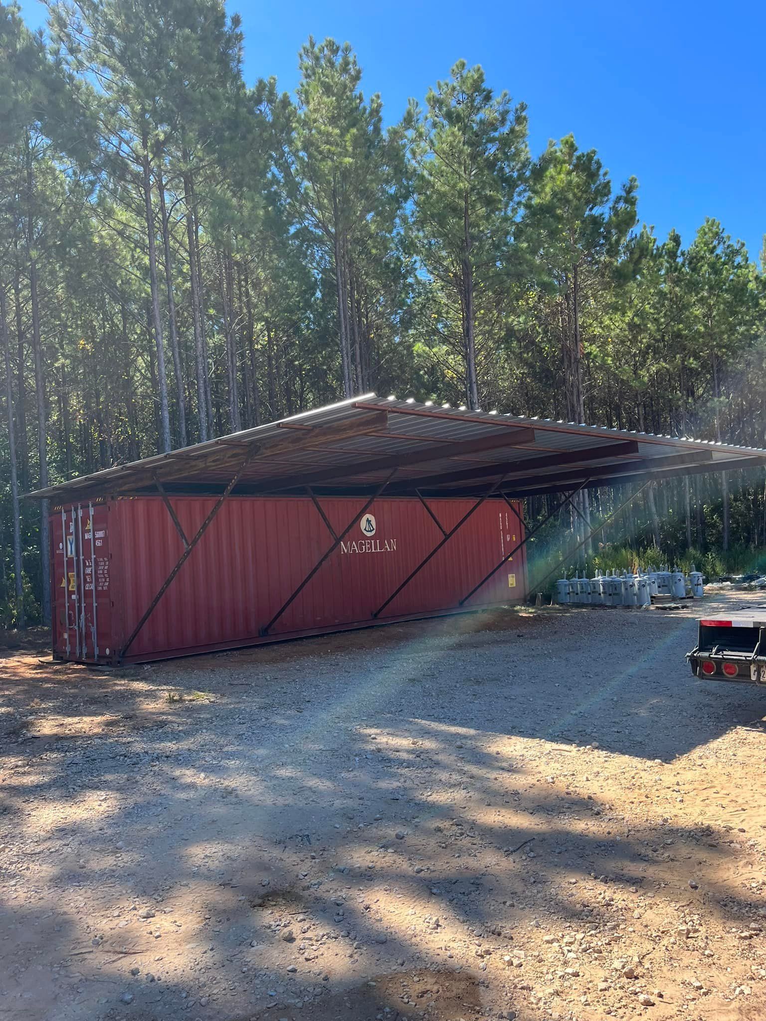 Red shipping container with a dark roof in a gravel area, trees in the background.