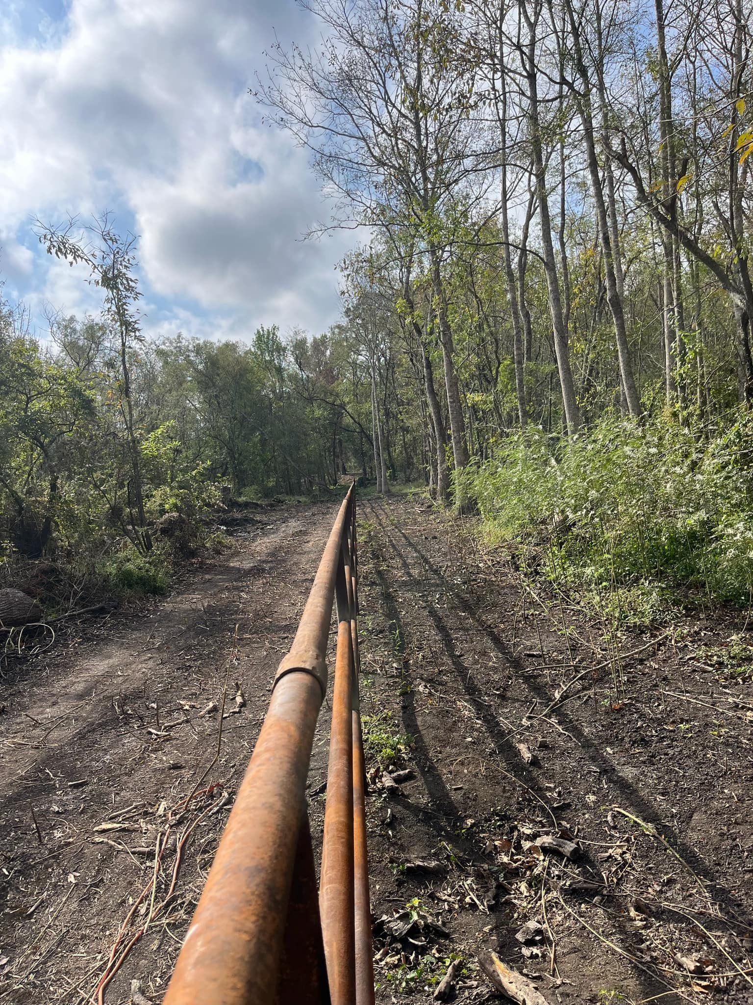 A rusty metal fence extends into a dirt path surrounded by trees under a cloudy sky.