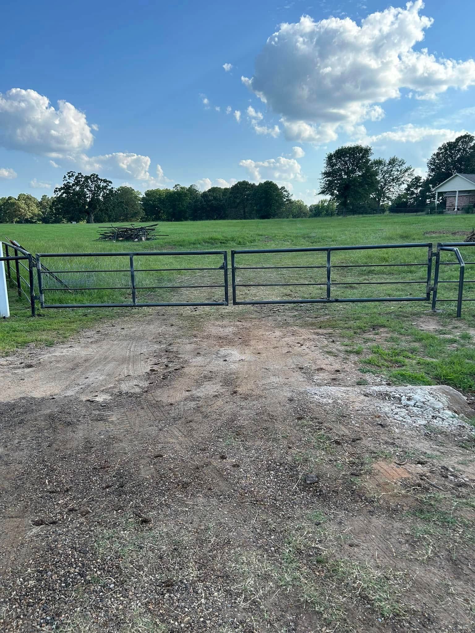 Metal gate on a dirt path leading to a green field under a blue sky with clouds.