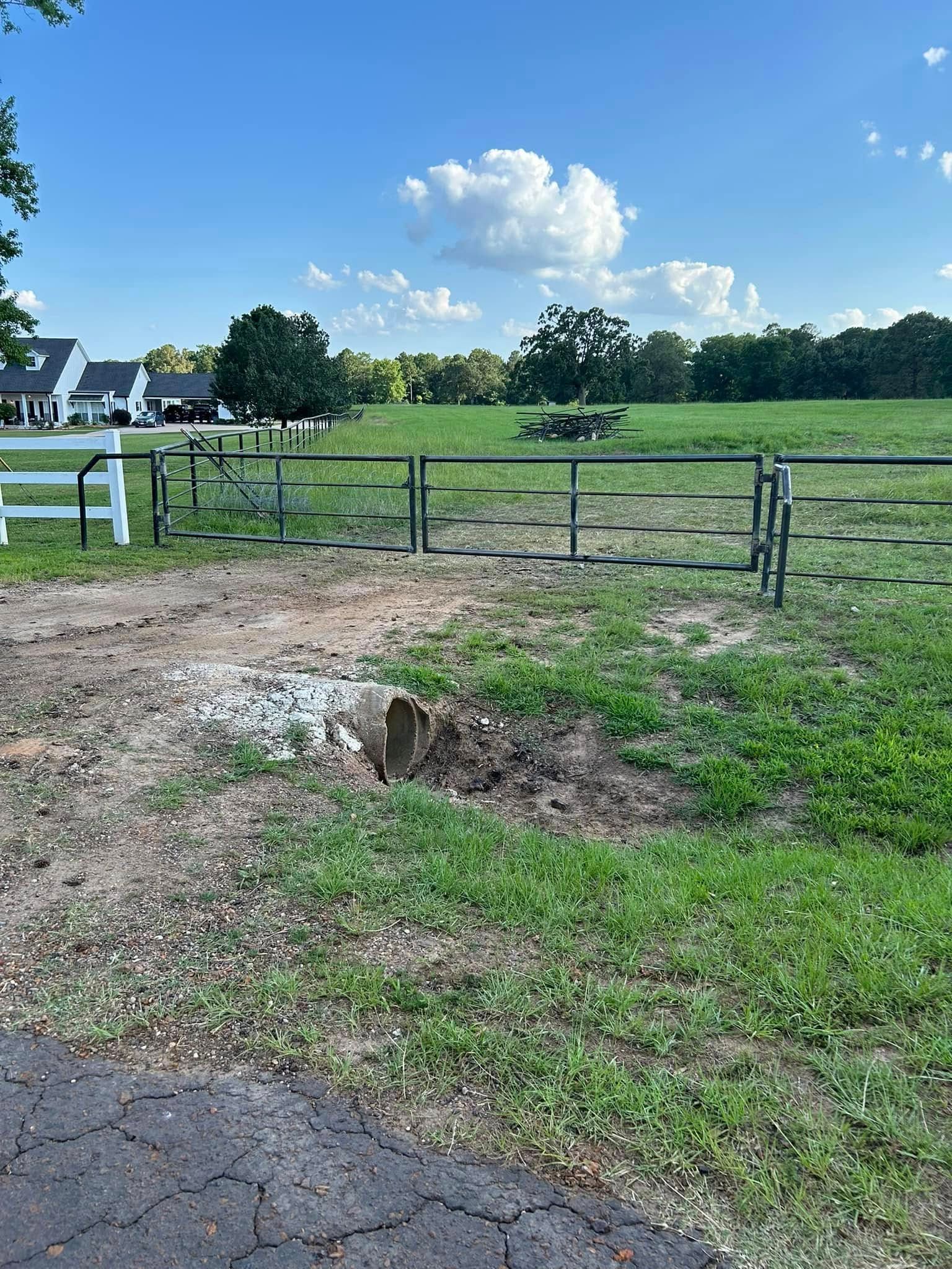 A field with metal gates, a grassy area, and a blue sky with clouds. Houses are visible in the background.