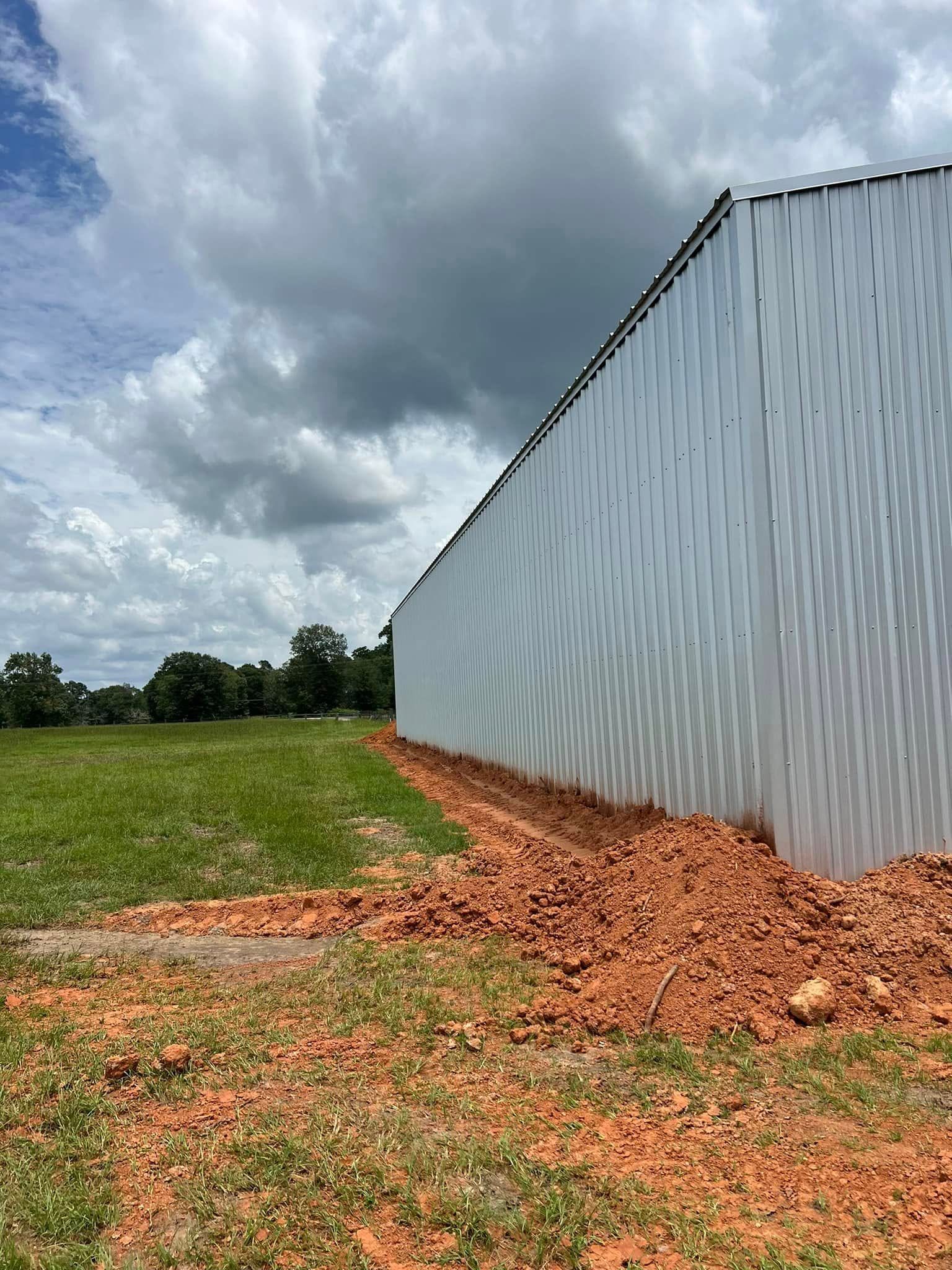 A white metal building with red clay soil next to it on a green field, under a cloudy sky.