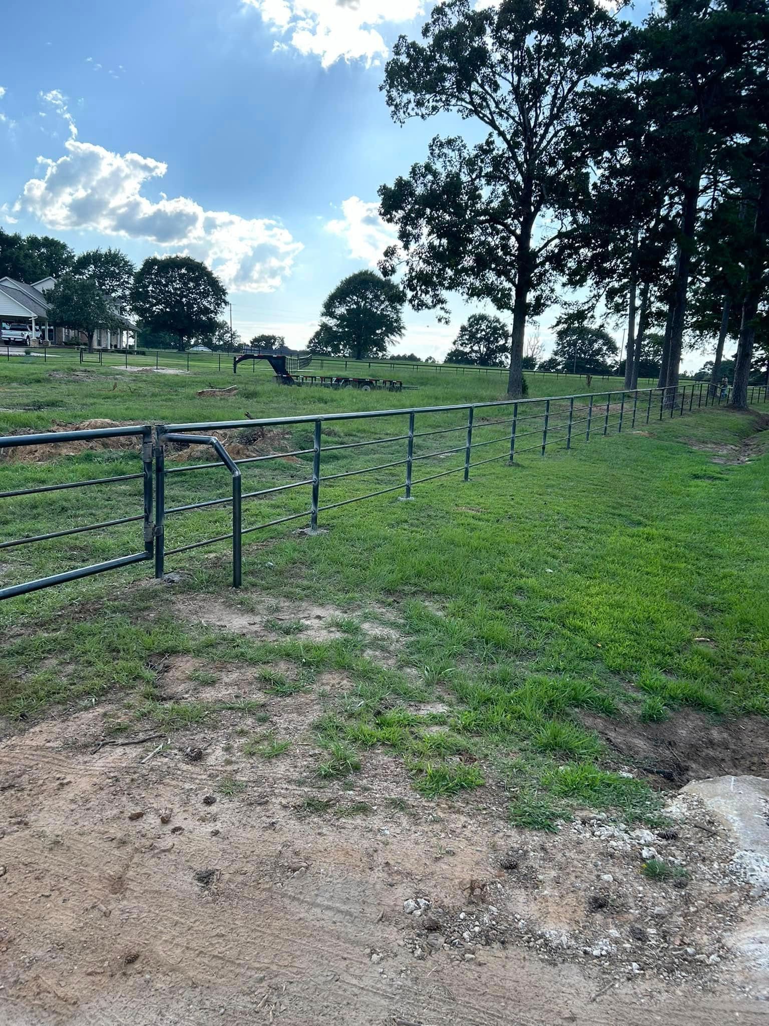 A green metal fence in a grassy field, with trees and a partly cloudy sky above.