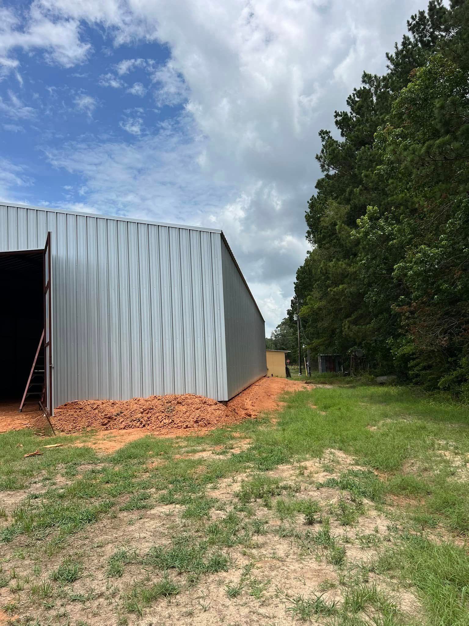 Metal building with patterned siding, surrounded by dirt, grass, and trees, under a cloudy sky.