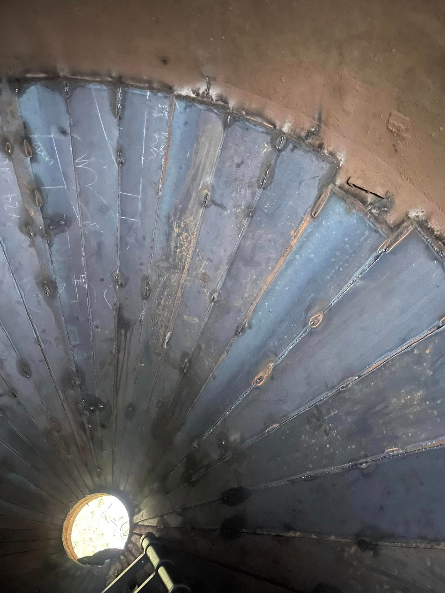 Inside of a metal silo, showing welded metal plates forming a conical shape leading to a round opening.