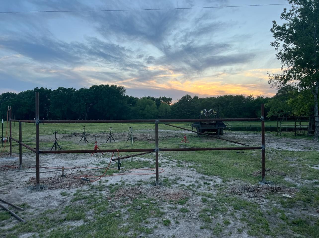 Metal fence under construction in a field, trees in background, sunset sky.