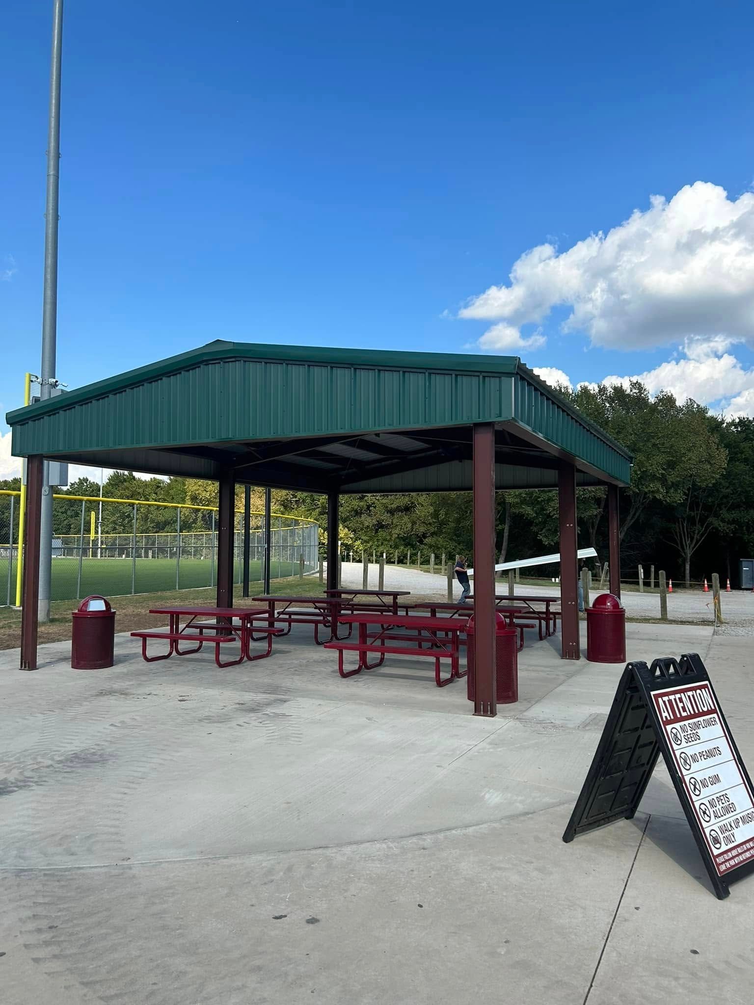Picnic shelter at park with tables, red benches, and trash cans. Green roof, brown columns, and a blue sky.