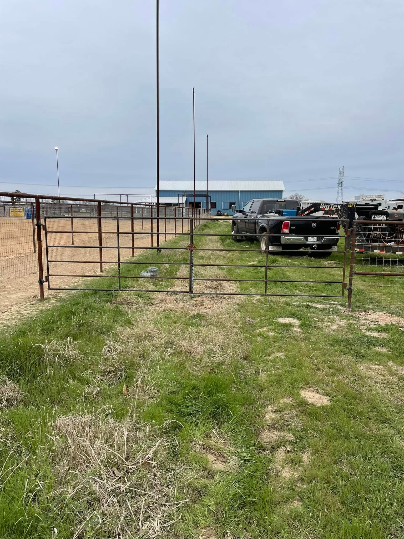 A metal fence and gate lead to a dirt area with vehicles and a blue building under a cloudy sky.