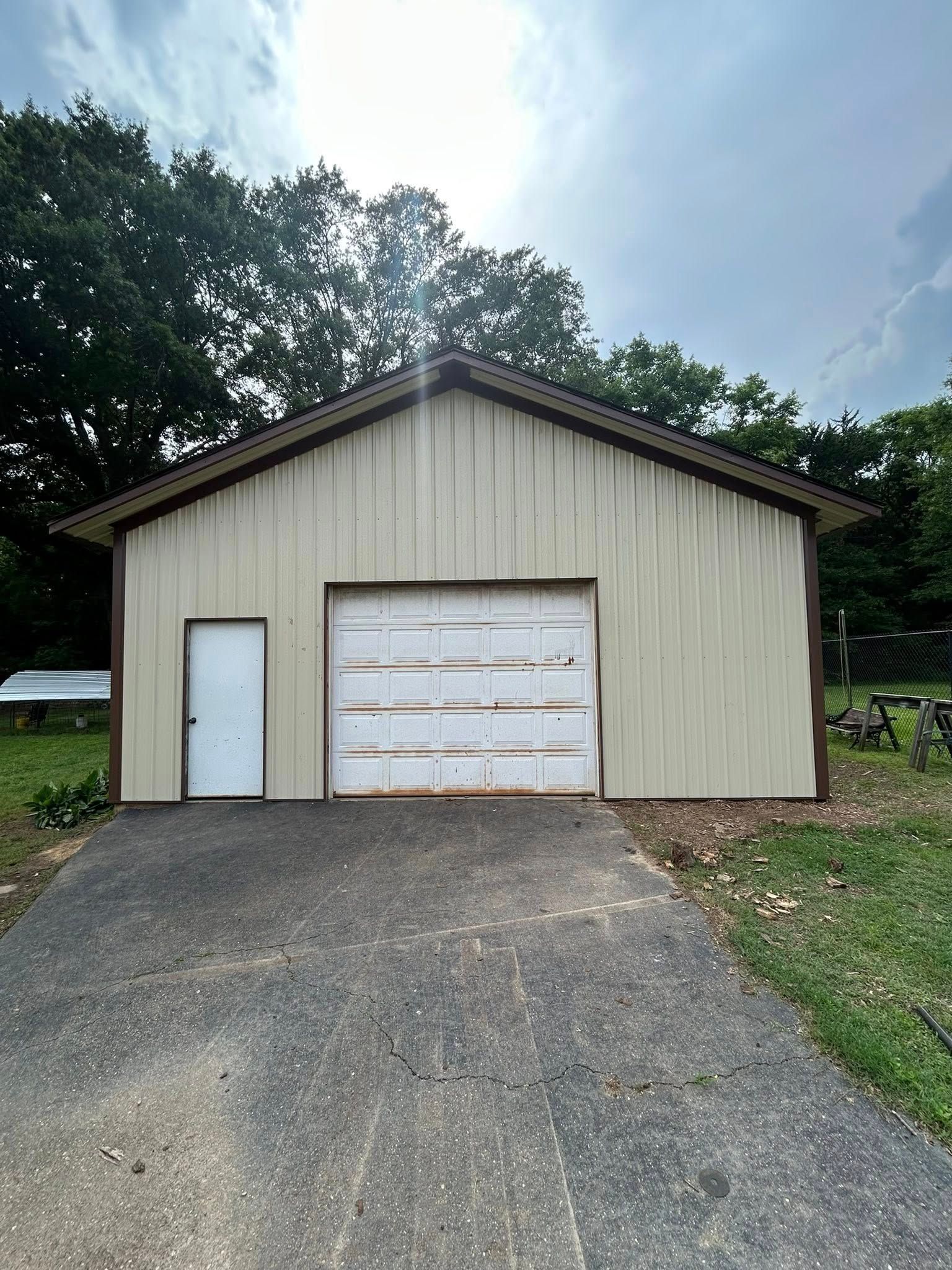 Tan metal garage with white door and driveway. Overcast sky and green trees in the background.