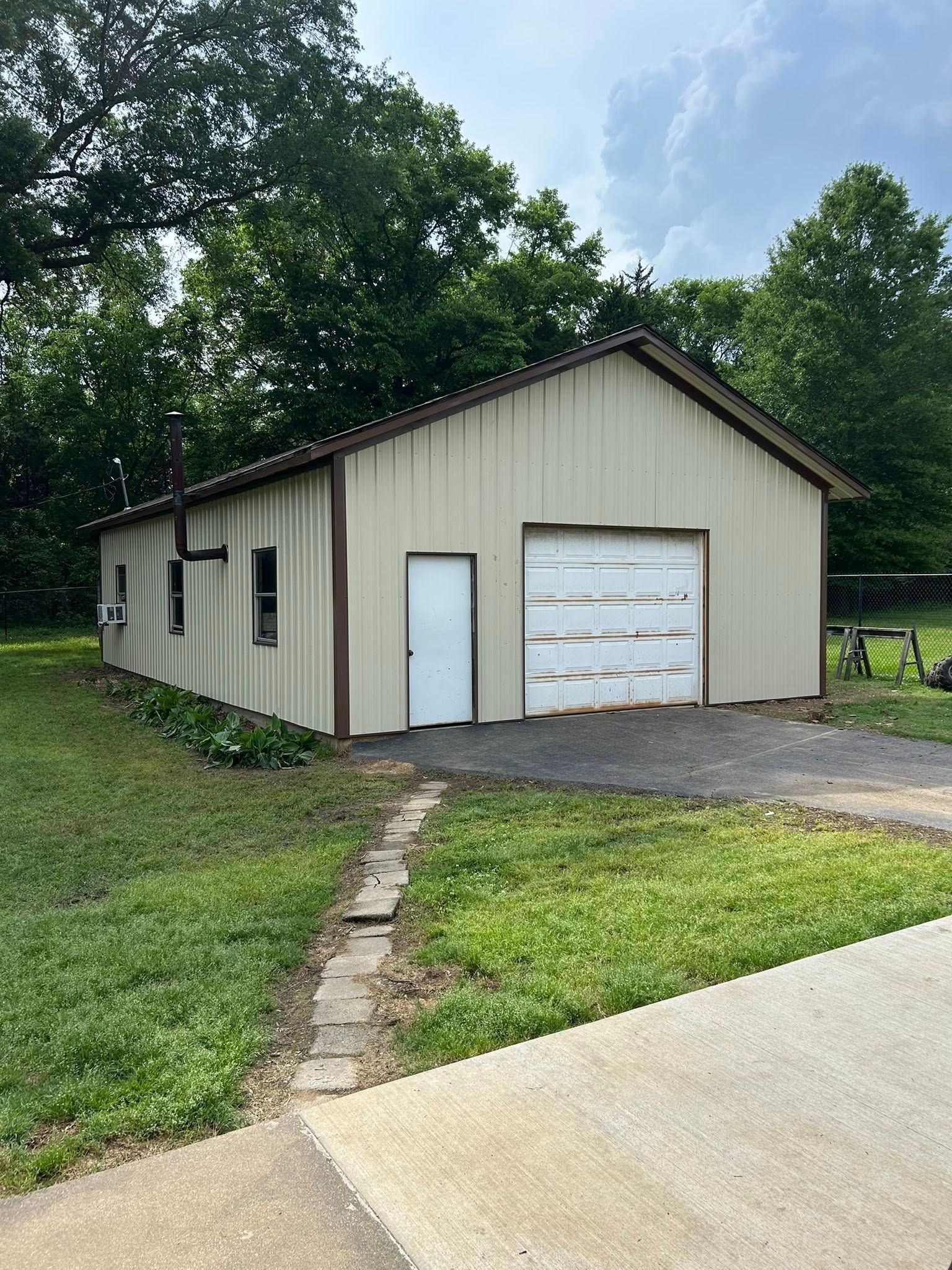 Beige metal garage with a white door and garage door, set in a grassy yard with trees.
