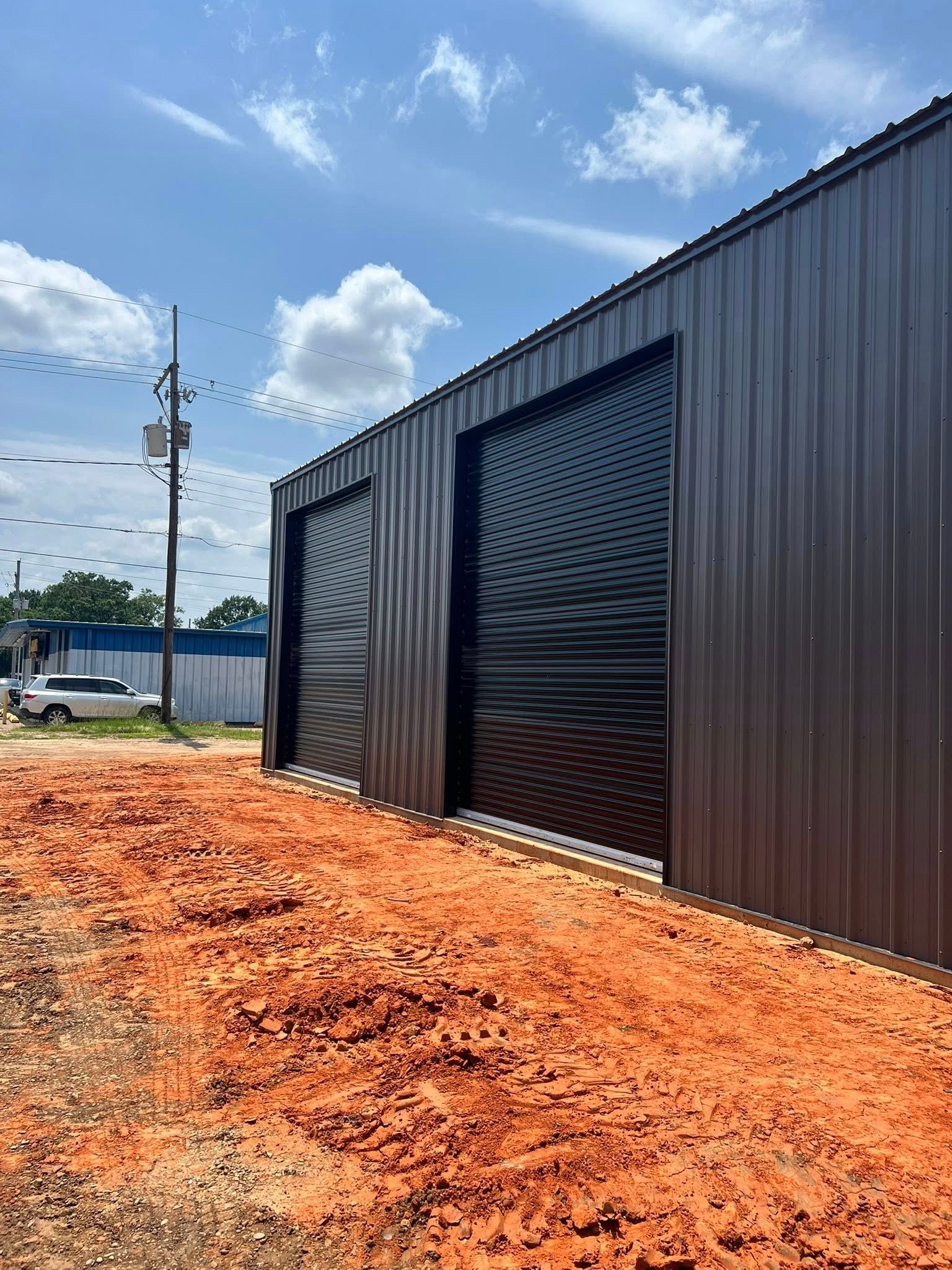 Brown metal building with black roll-up doors, in front of reddish-brown dirt, under a cloudy blue sky.