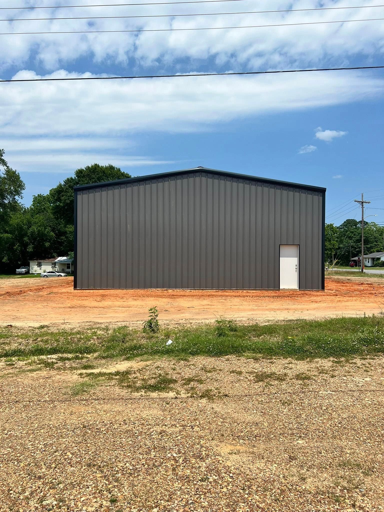 Gray metal building with a white door, on a dirt lot with blue sky and some trees in the background.