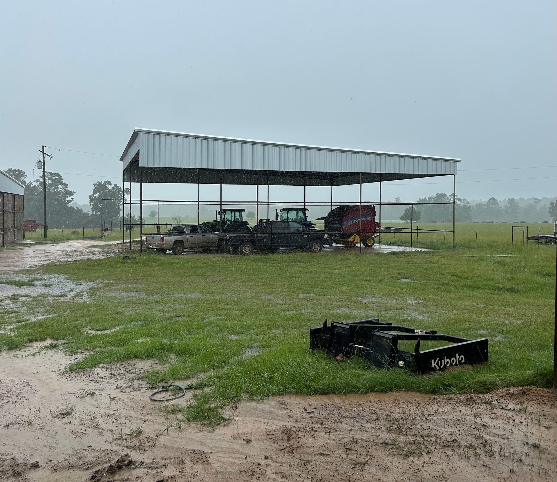 A rural scene in the rain. Farm equipment parked under a metal shelter on a muddy field.