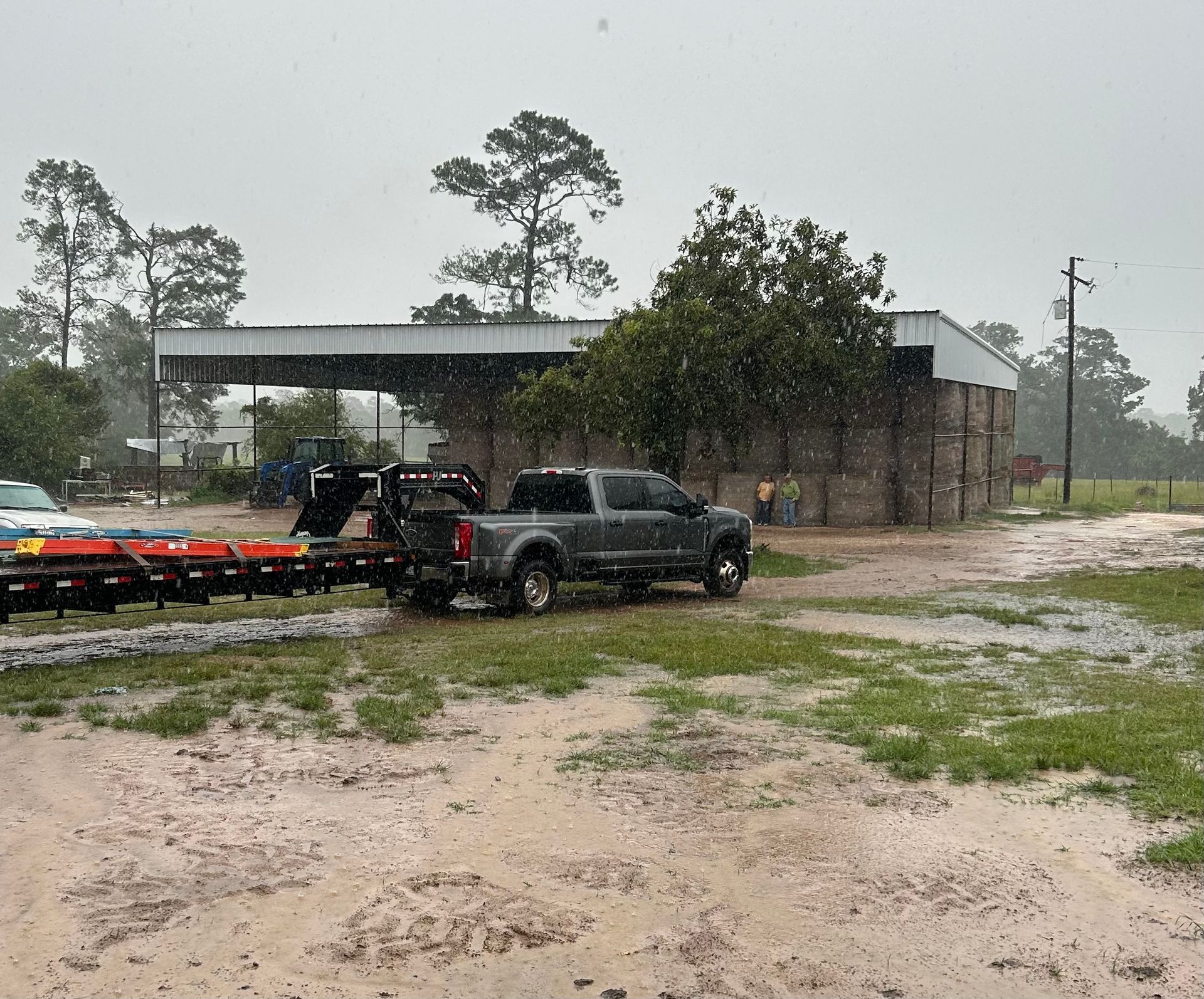 Truck with trailer in front of a barn in the rain. Muddy ground, bales of hay stacked nearby.