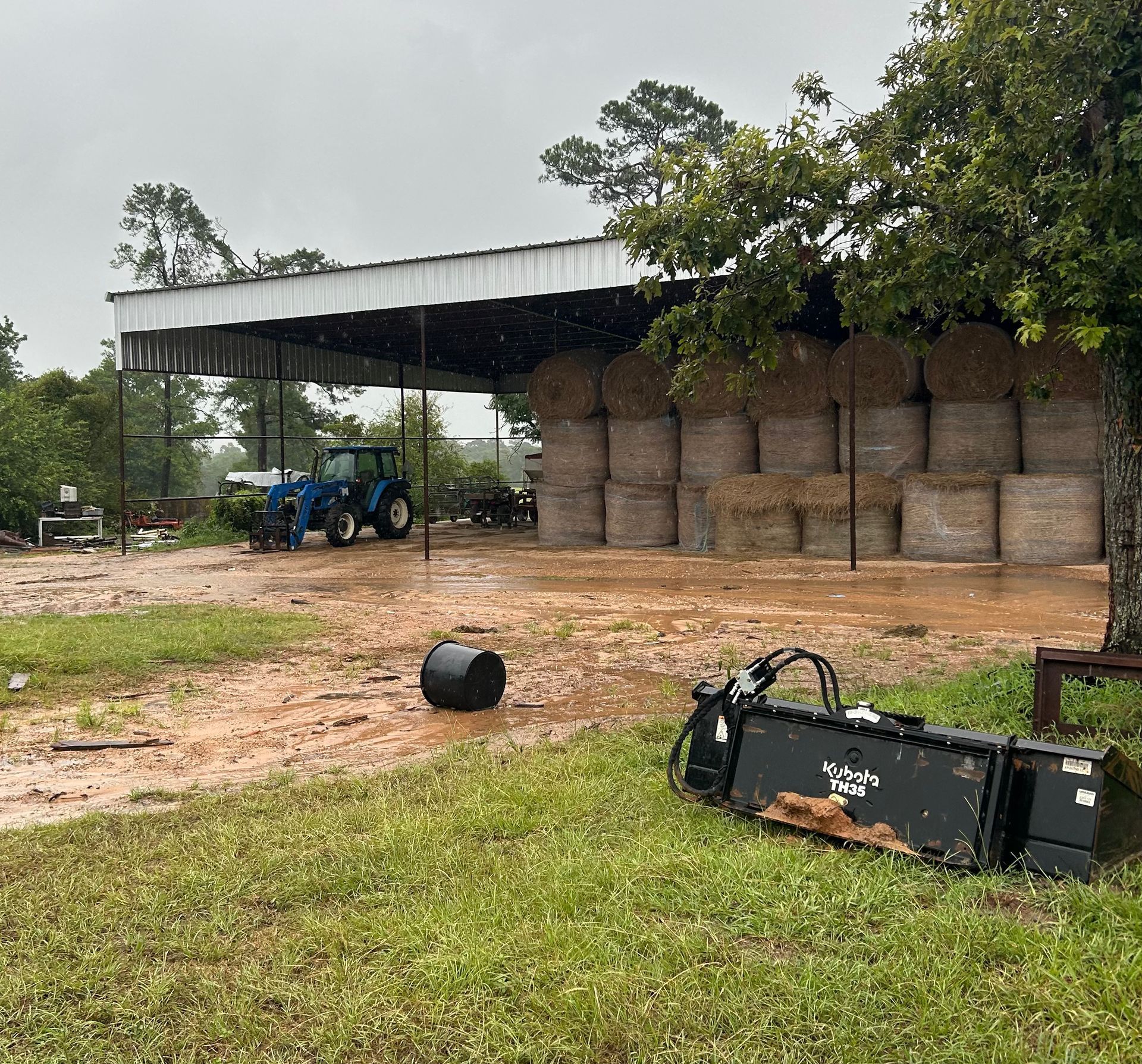 Muddy farm scene with a tractor, hay bales stacked under a shed, and a front loader. Rain is falling.