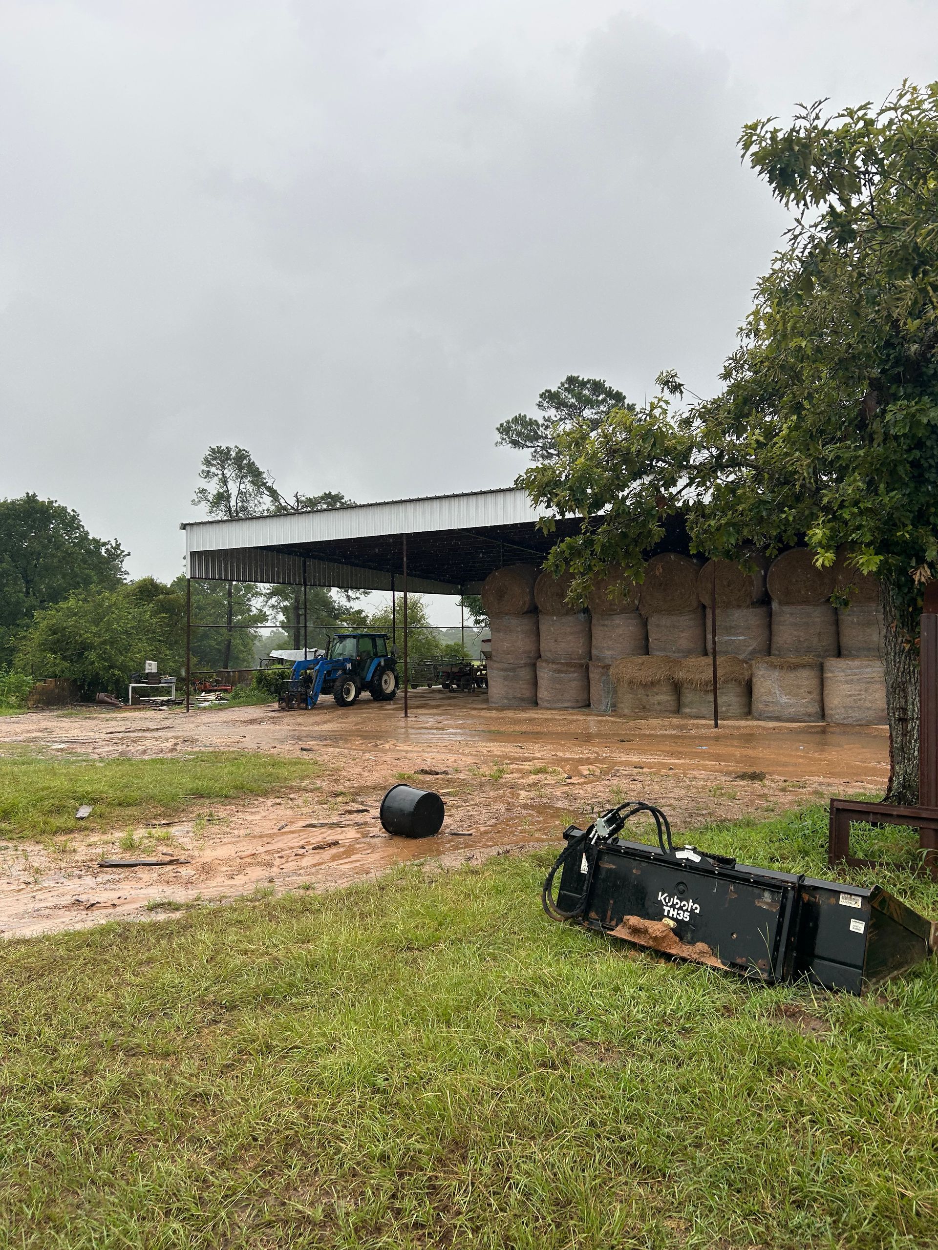 Farm scene: hay bales stacked under a shed, tractor nearby, loader bucket in foreground, overcast.