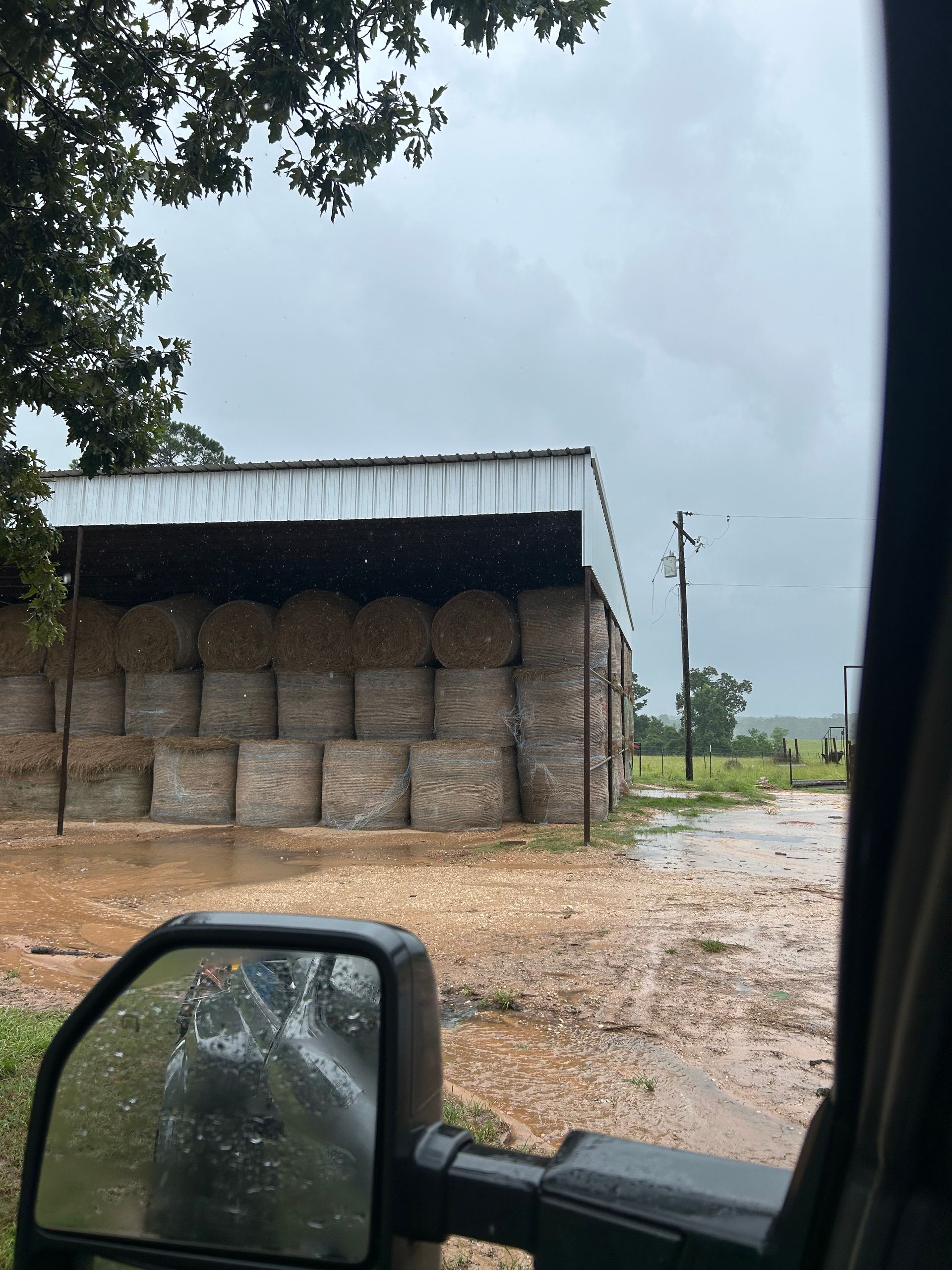 Rainy day view: stacked hay bales under a metal roof, waterlogged ground, seen from inside a vehicle.