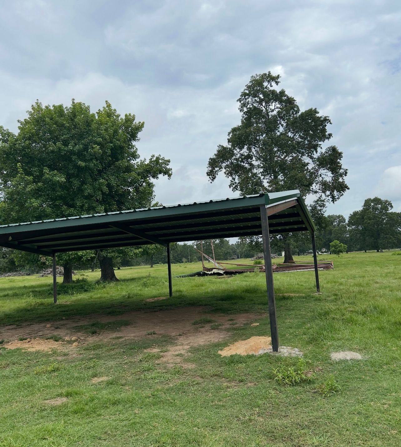 Open-sided metal shelter in a grassy field with trees under a cloudy sky.