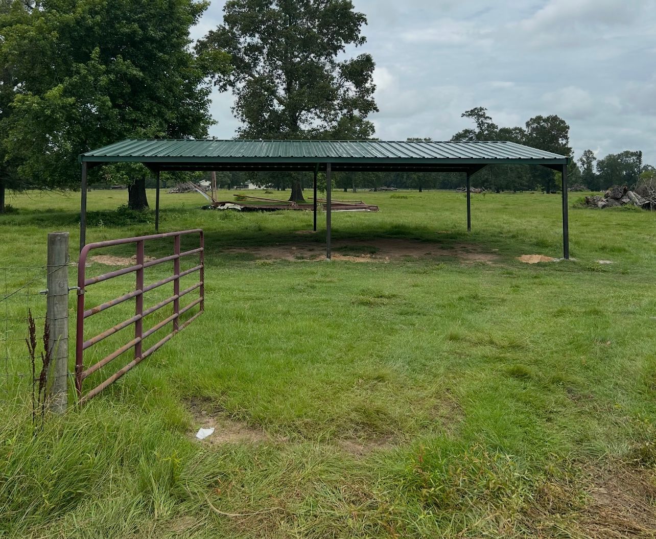 Open metal shelter in a grassy field with a red gate and a tree in the background.