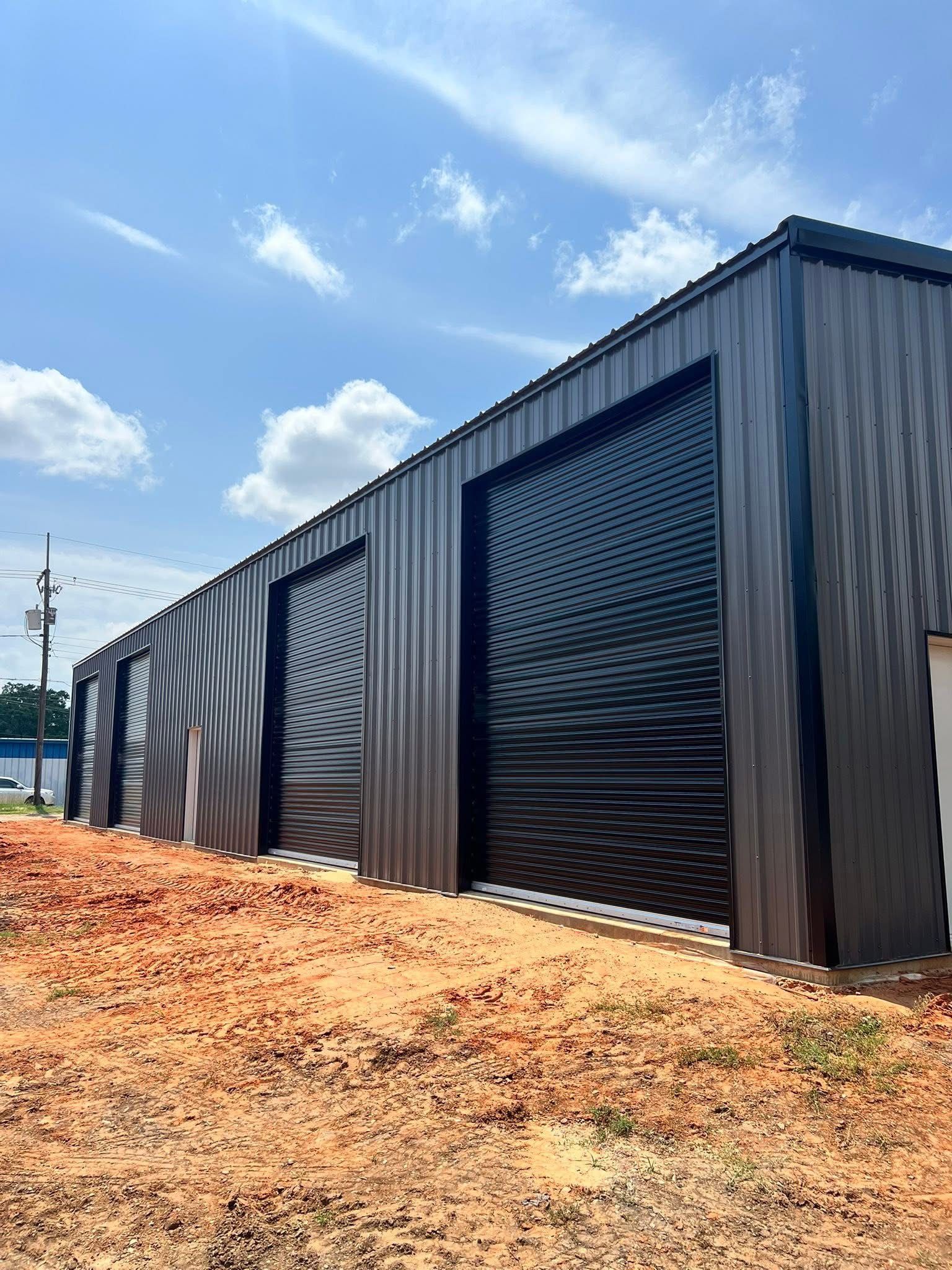 Dark metal building with three large roll-up doors, under a blue sky with clouds.