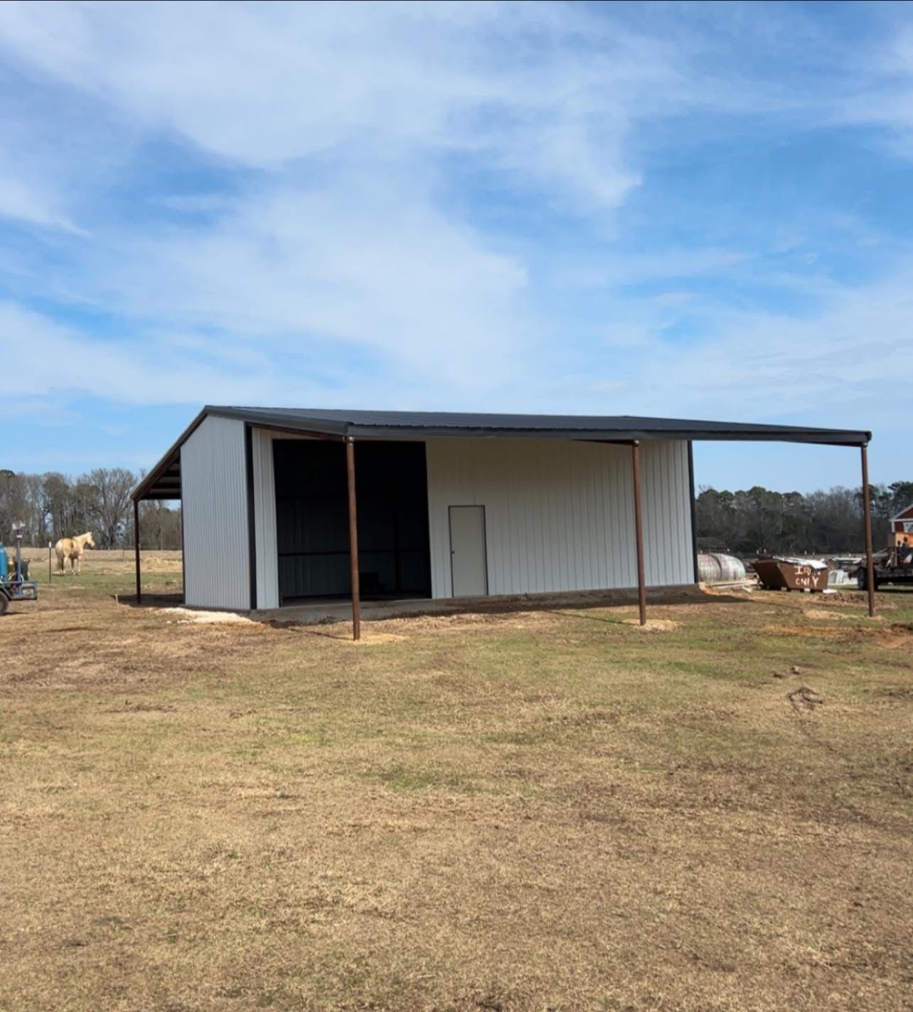 A white metal barn with a dark roof and open-air awning on a grassy field under a blue sky.