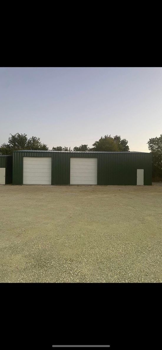 Green metal storage building with three large doors on a gravel lot with a clear blue sky.