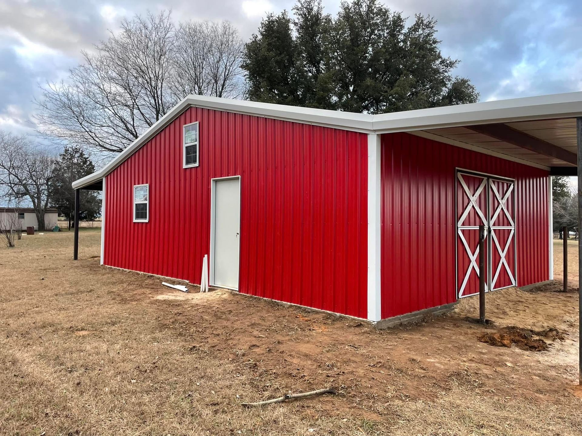 Red metal barn with white trim and open-air carport.  Two small windows, door. Brown field.