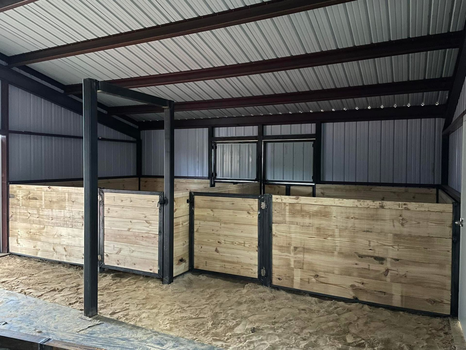Inside of a horse stable with stalls, light brown wood walls, gray steel structure, and dirt floor.