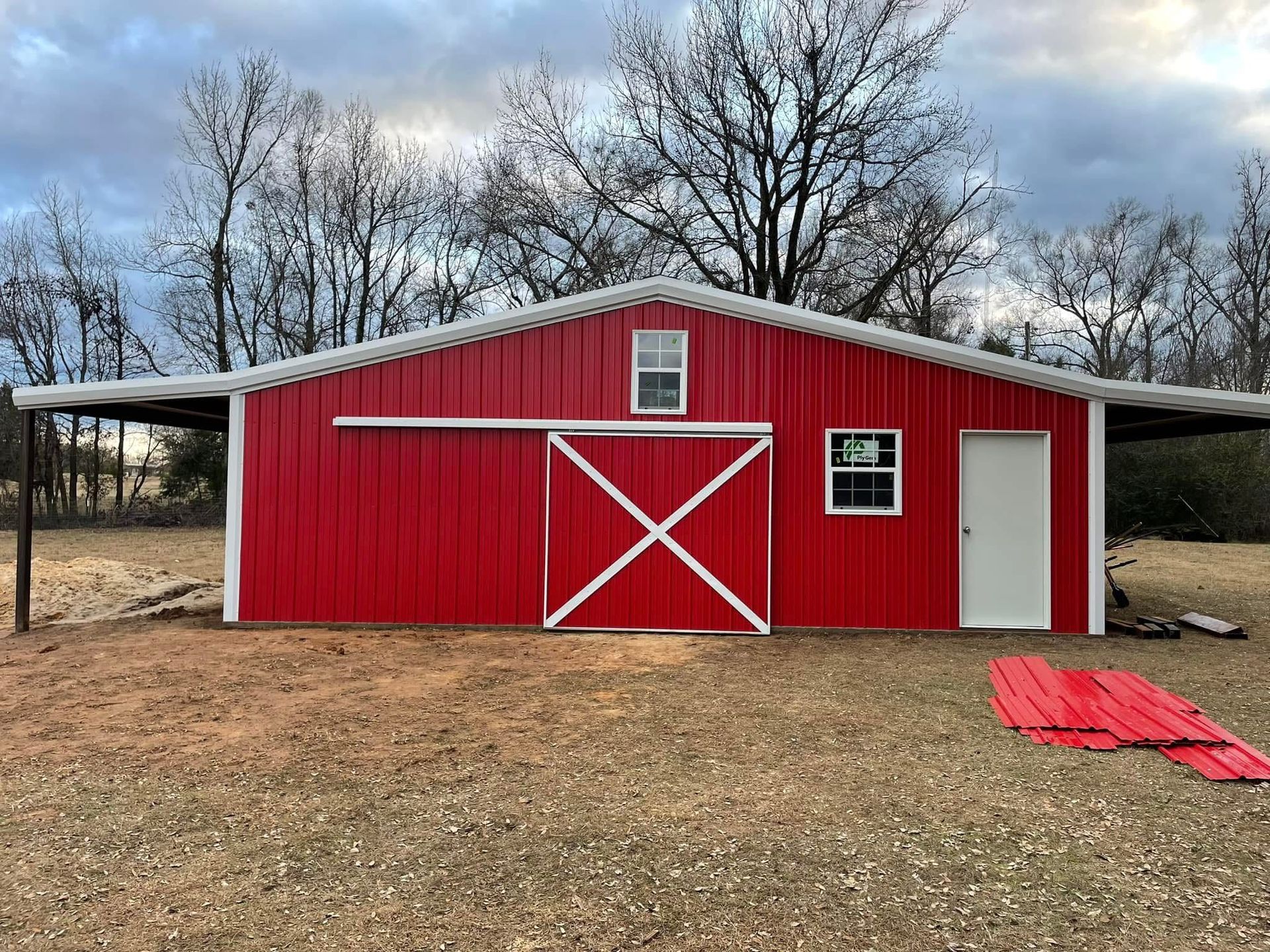 Red barn with white trim and sliding door, flanked by covered areas; set on a grassy field with trees.