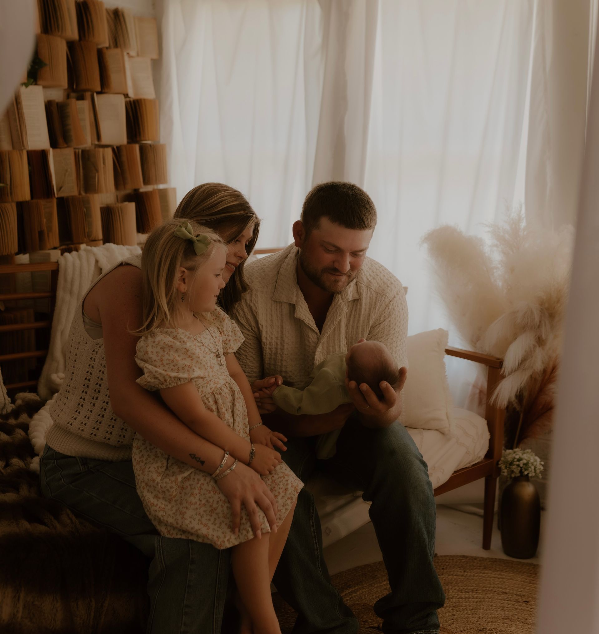 Family of four sits together, gazing at a newborn. Sunlight streams through curtains.