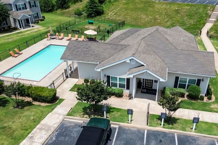 Aerial view: apartment clubhouse, pool, and dog park. Building is light with dark shutters; pool has lounge chairs.