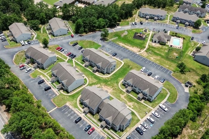 Aerial view of apartment complex with multiple buildings, parking, pool, and surrounding trees.