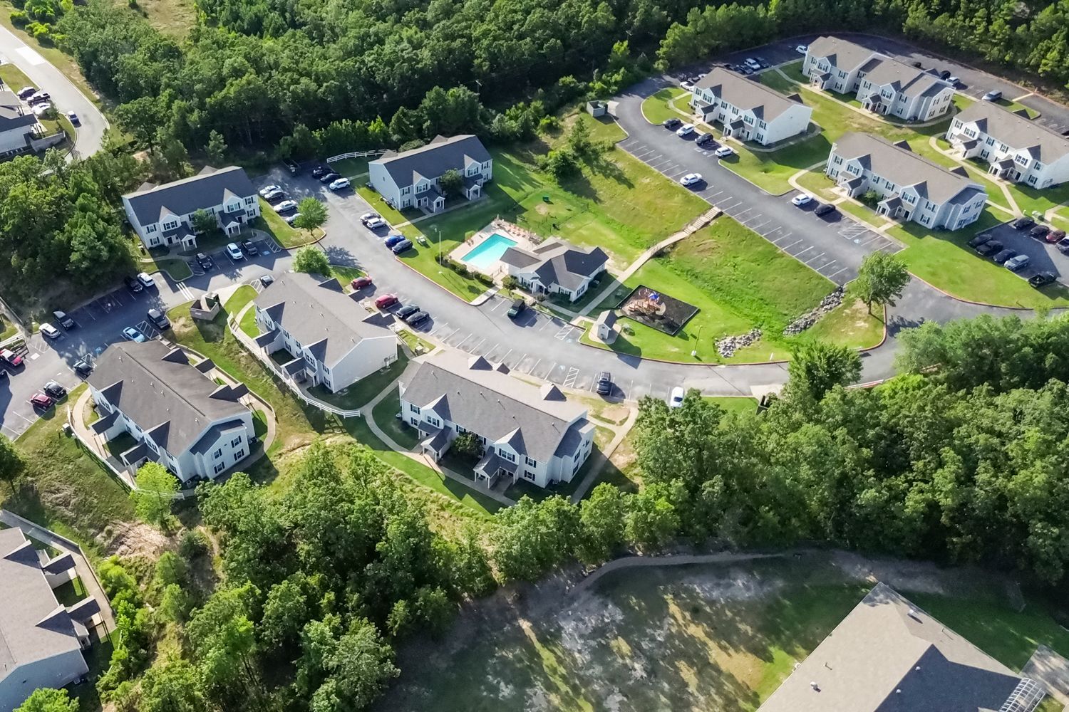 Aerial view of apartment complex surrounded by trees, with a swimming pool and parked cars.