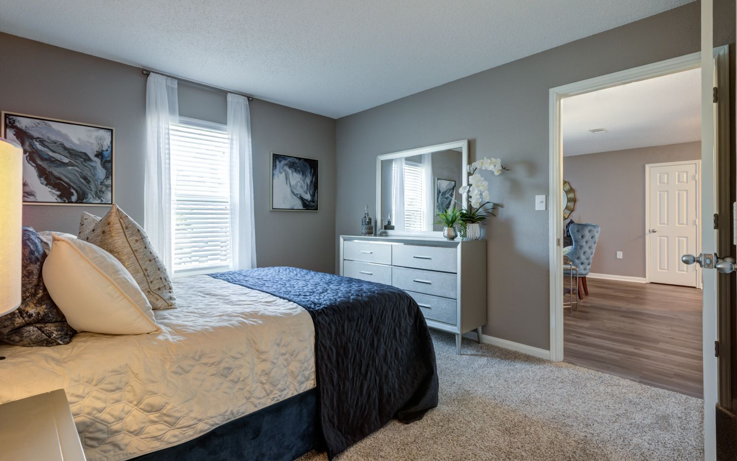 Bedroom with bed, dresser, art, window, and open door to another room. Gray walls, white trim, and carpet.
