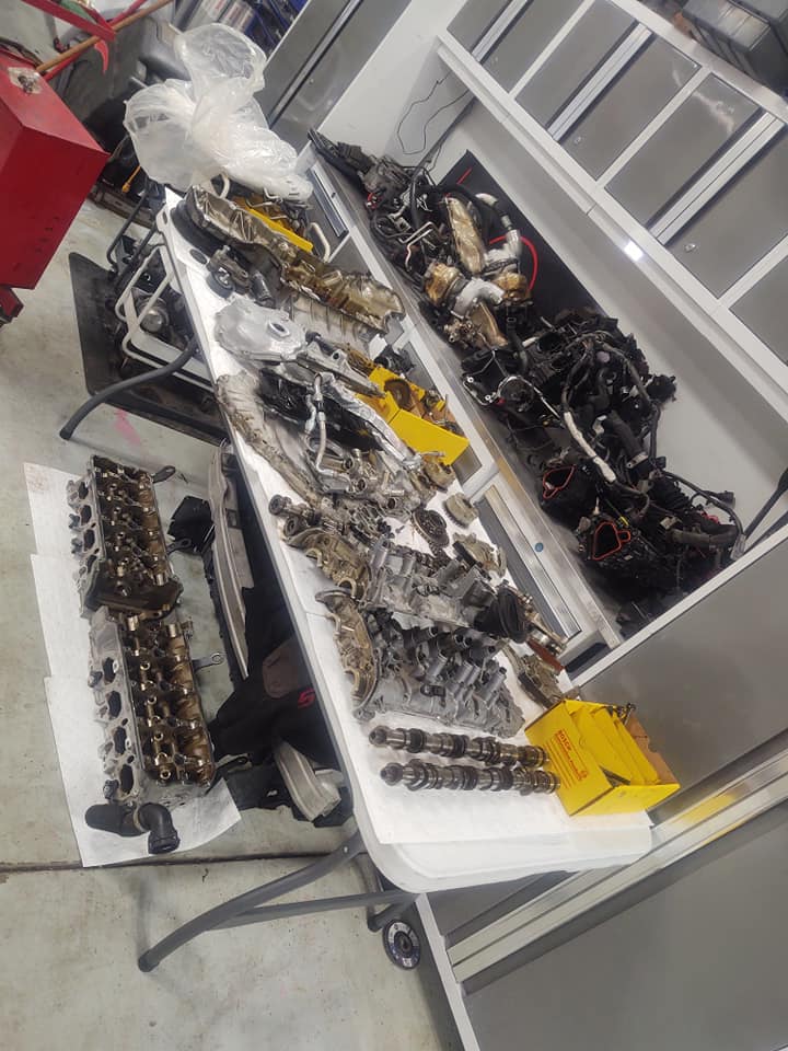 Disassembled engine parts spread on a white table in a garage. Includes cylinder heads, belts, and other components.