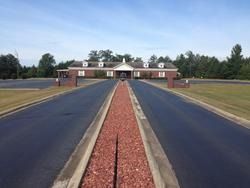 Long driveway leads to a brick building with a central portico, red rock median, and trees under a blue sky.
