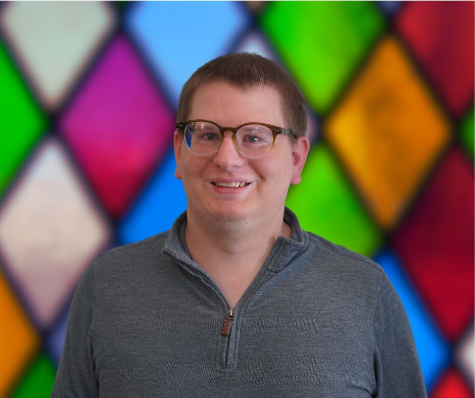 A man wearing glasses and a blue shirt is standing in front of a stained glass window.