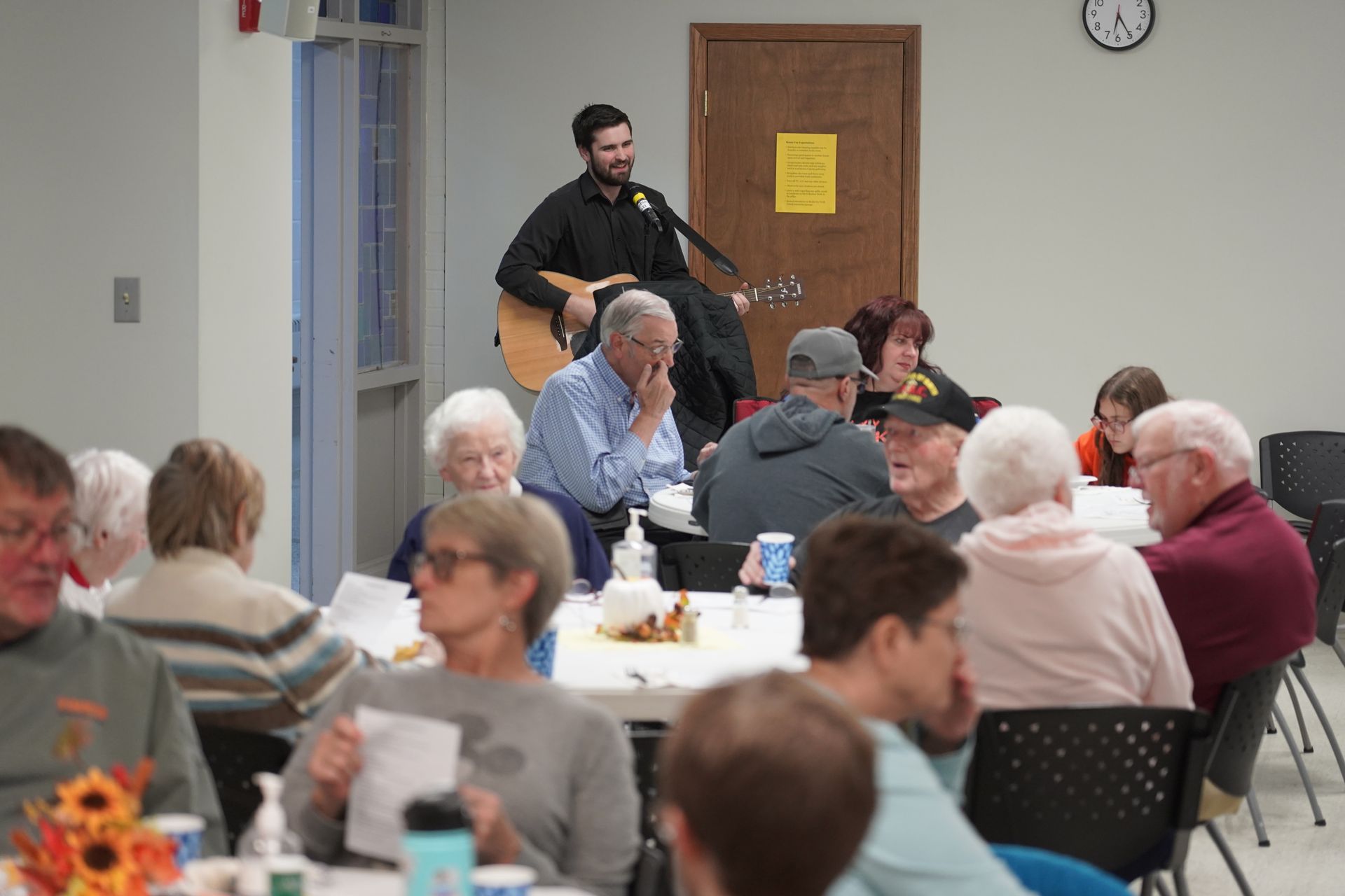 A man is playing a guitar in front of a group of people sitting at tables.