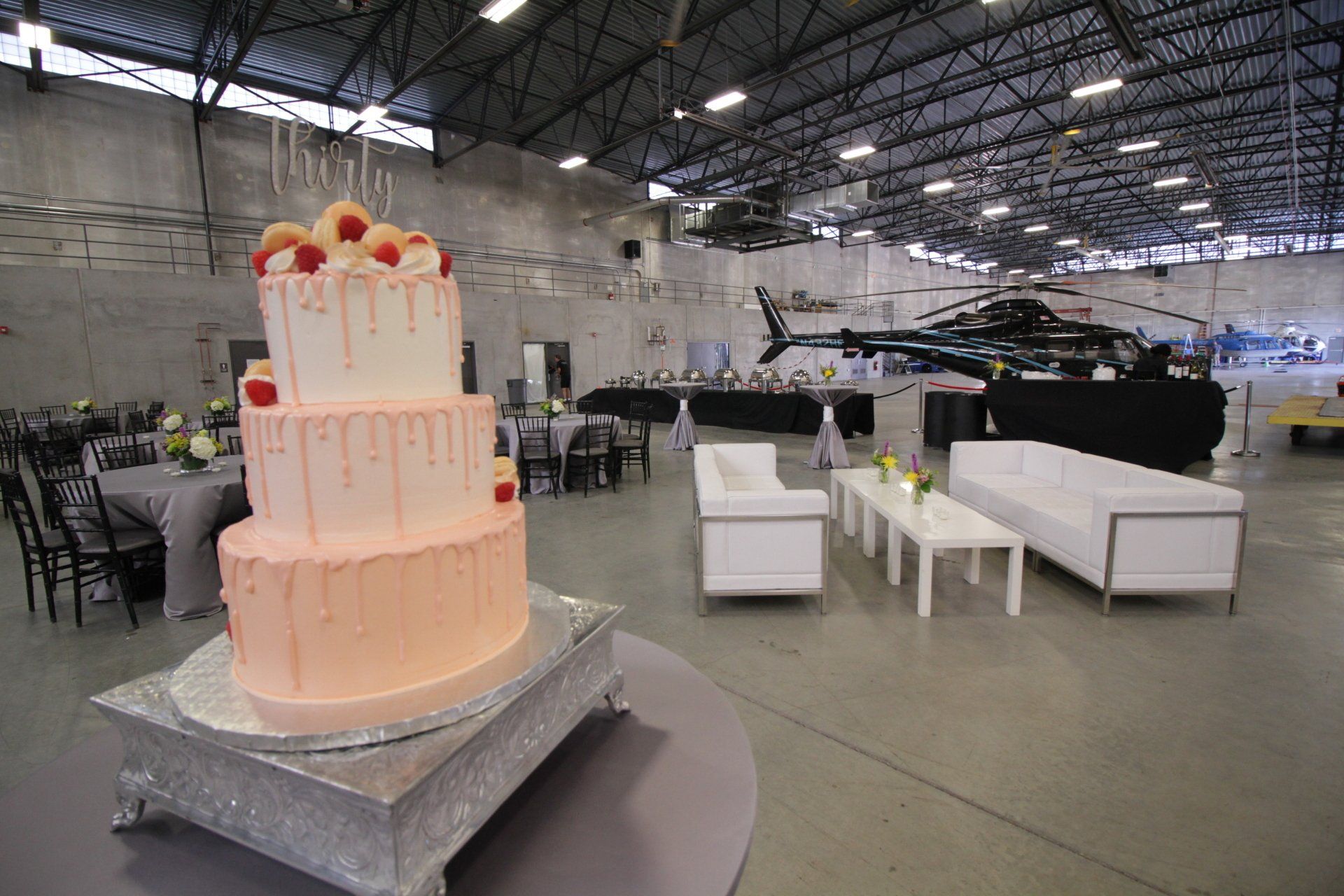 A large pink cake is sitting on top of a silver cake stand in a large room.