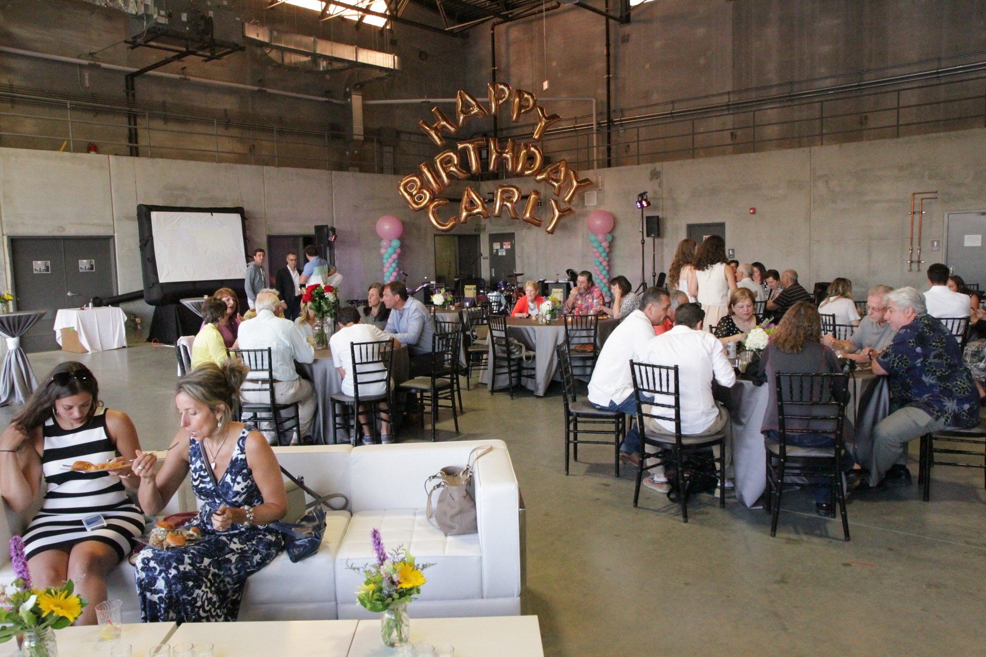 A large group of people are sitting at tables in a room with balloons that say happy birthday carly.