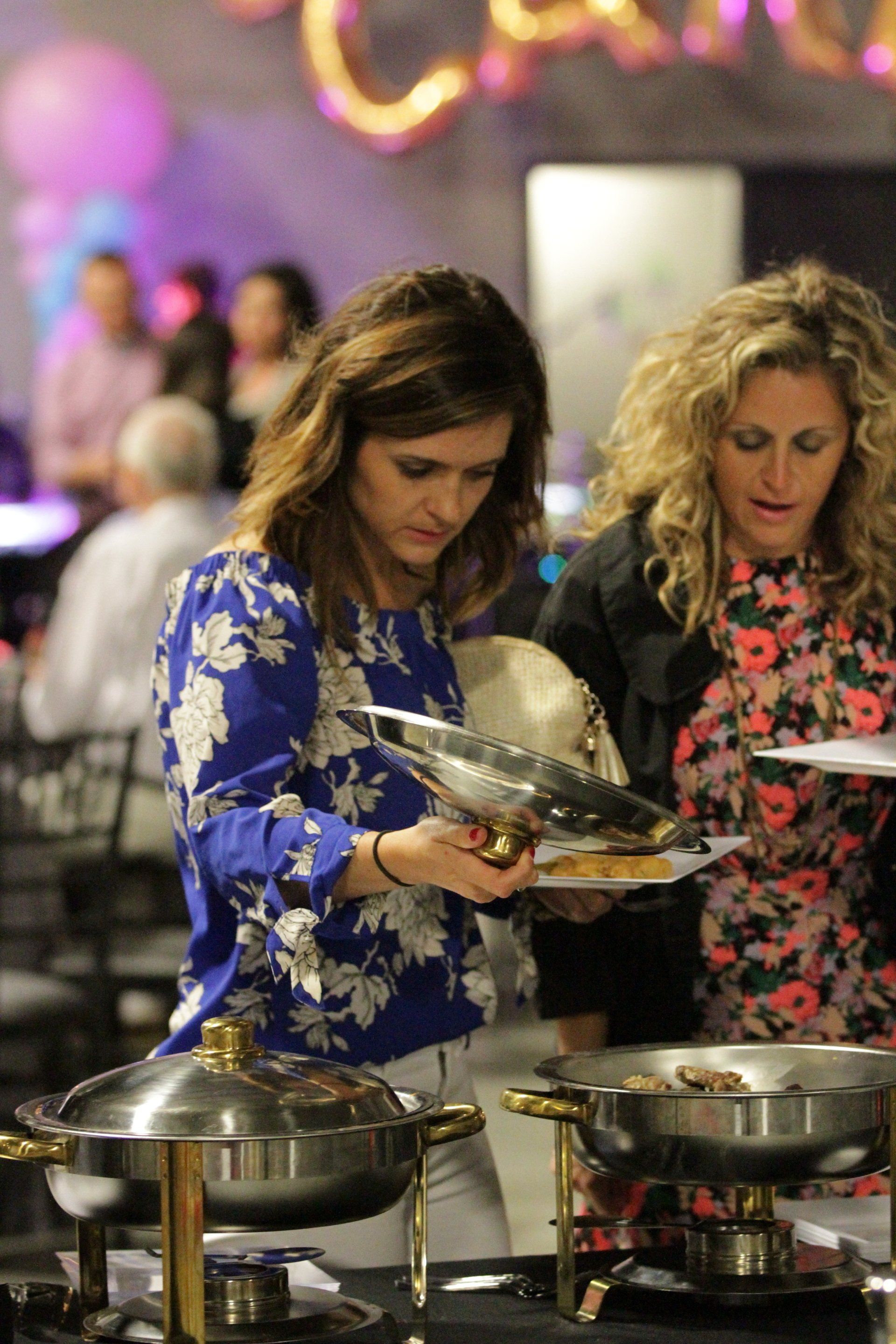 Two women are standing in front of a buffet line at a party.