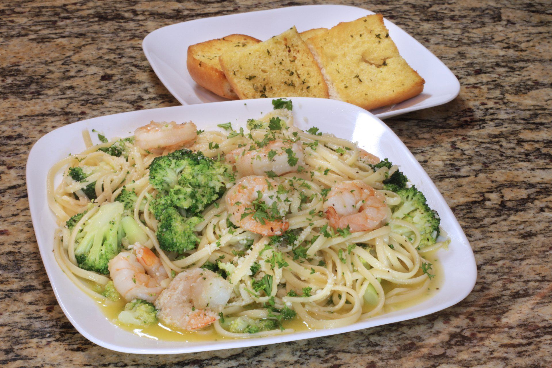 A plate of spaghetti with broccoli and shrimp next to a plate of garlic bread