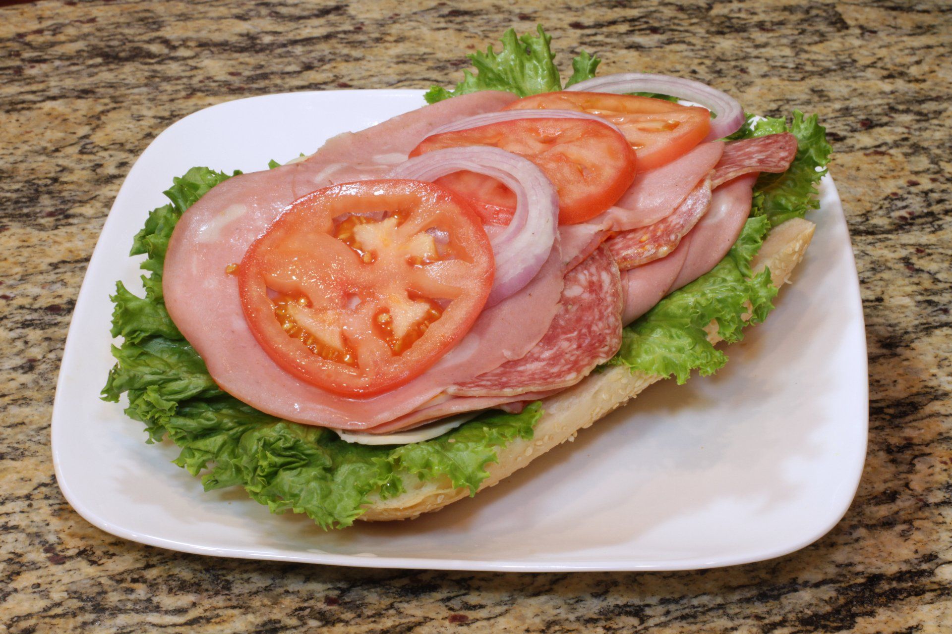 A sandwich with ham , lettuce , tomatoes and onions on a white plate.