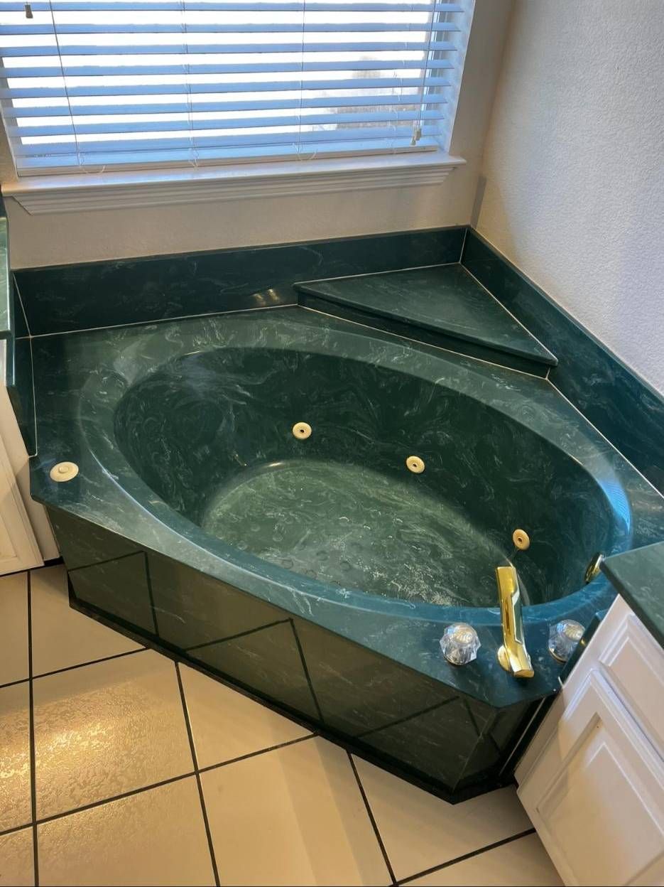 A corner-fitted jetted bathtub made of dark green marble with a gold-tone faucet, set against a window with white blinds.