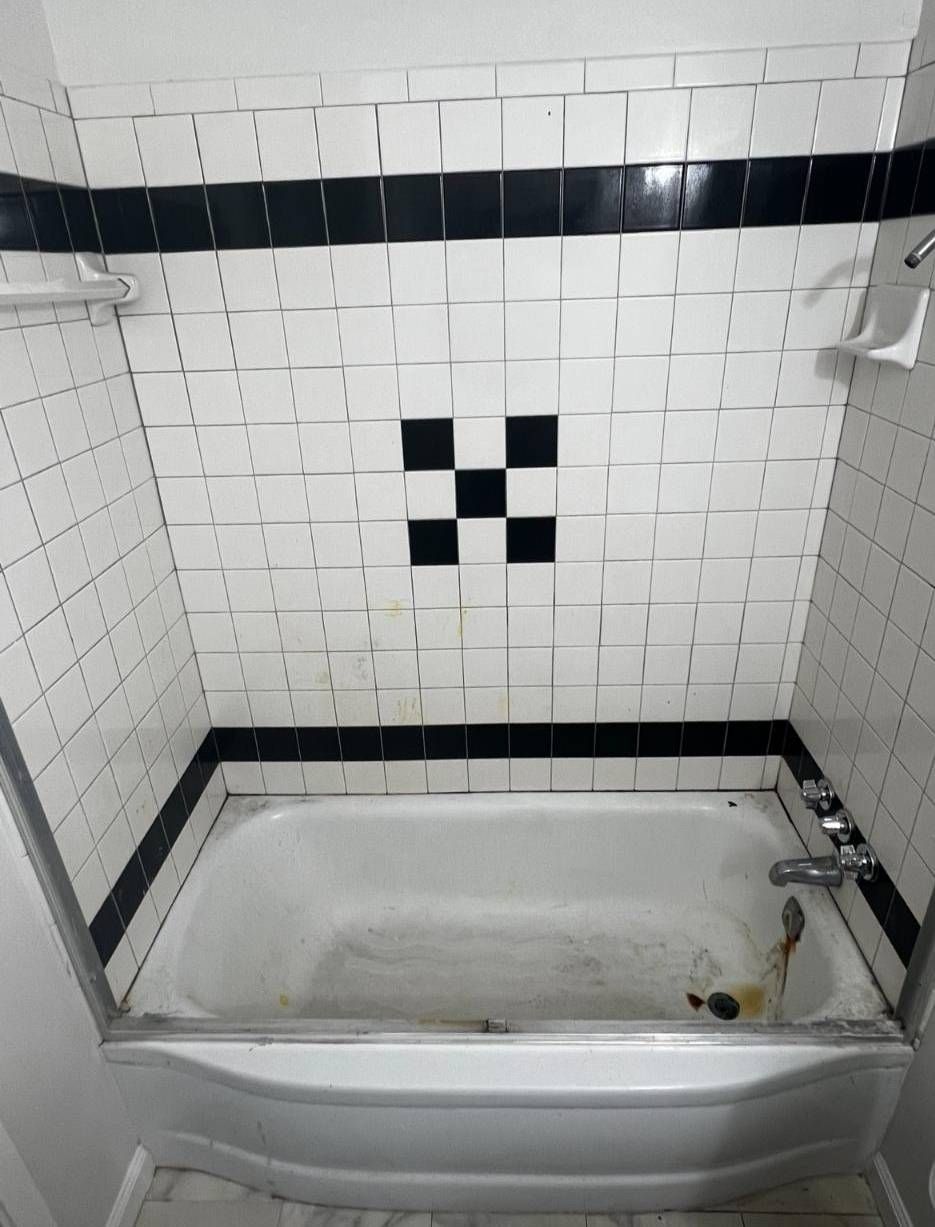 A bathtub in a shower enclosure with white tiled walls featuring black accents and a small black cross-shaped design.
